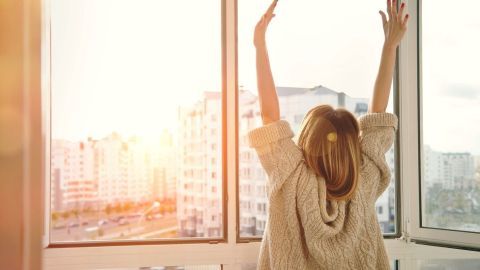Eine Person steht vor einem Fenster, reckt die Arme nach oben und genießt das Licht des Morgen, was ein Gefühl von Wachheit vermittelt.