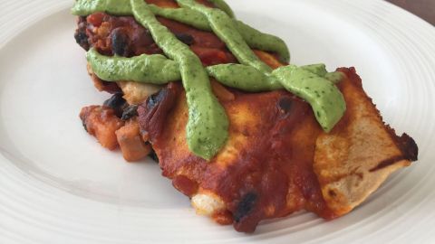 Eine Enchilada mit Süßkartoffeln und schwarzen Bohnen liegt auf einem weißen Teller, garniert mit grüner Soße.