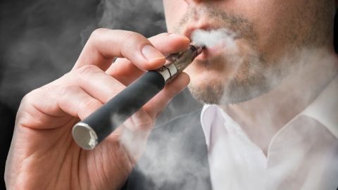Ein Mann raucht eine E-Zigarette, während Dampf in einem dunklen Raum aufsteigt. E-Zigaretten sind umstritten und ungesund.