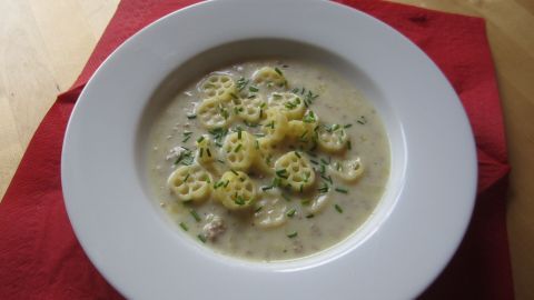 Eine cremige Käse-Lauch-Suppe mit Hackfleisch und knusprigen Kartoffelrösti, angerichtet in einer weißen Schüssel auf rotem Untergrund.