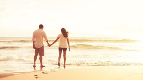 Ein Paar hält Händchen, während es am Strand entlangschlendert. Die Wellen brechen sanft im Hintergrund, Sonnenlicht umhüllt sie.