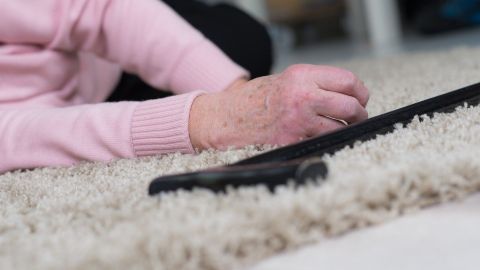 Eine Hand liegt auf einem Teppich, während ein Gehstock daneben liegt. Die Umgebung wirkt ruhig und häuslich.