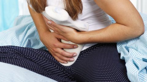 Eine Frau sitzt auf einem Bett und hält sich einen Rollwagen gegen den Unterbauch, um die Beschwerden einer Blasenentzündung zu lindern.