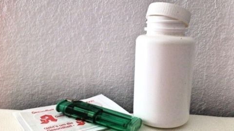 Ein weißes Fläschchen steht neben einer grünen Spritze und einem kleinen Paket auf einer flachen Oberfläche.