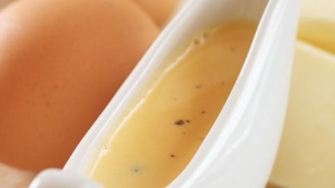 Eine weiße Sauciere mit Sauce Hollandaise steht in der Mitte. Im Hintergrund liegen zwei Eier und ein Zitronenstück.