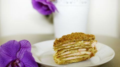 Eine Pfannkuchentorte mit Himbeercreme steht auf einem weißen Teller. Daneben liegt eine lila Blume, im Hintergrund ein Pflanzentopf.