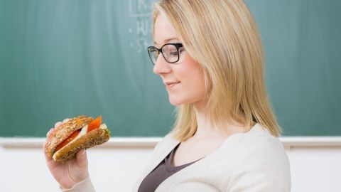 Eine blonde Frau mit Brille hält ein Sandwich in der Hand und schaut es nachdenklich vor einer grünen Tafel an.
