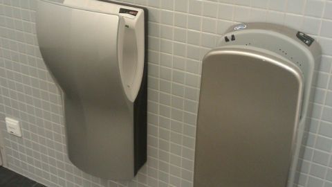 Ein Händetrockner aus Edelstahl bläst Luft zur Trocknung von Händen in einem modernen, gefliesten Badezimmer.