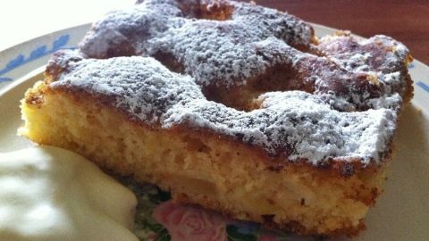 Ein saftiges Stück Apfelkuchen vom Blech liegt auf einem dekorativen Teller, neben einer Portion Sahne.