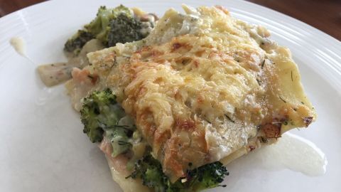 Eine cremige Brokkoli-Lachs-Lasagne liegt auf einem weißen Teller, mit goldbrauner Käsekruste und frischem Gemüse.