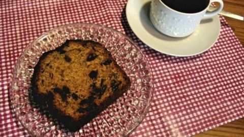 Ein Bananen-Karotten-Cranberry-Kuchen liegt auf einem gläsernen Teller neben einer Tasse Kaffee auf einem karierter Unterlage.