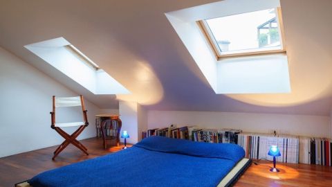 Ein blaues Bett liegt auf einem Holzfußboden, während Dachfenster Licht in den Raum lassen und vor Hitze schützen.