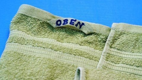 Ein Handtuch ruht auf einem blauen Hintergrund, zeigt den gestickten Text „OBEN“ und symbolisiert Hygiene und Sauberkeit.