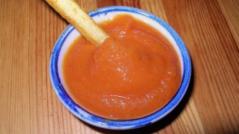 Eine Schale mit Karottenketchup steht auf einem Holztisch, während ein Stück Brot darin eingetaucht wird.