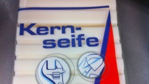 Kernseifenstück liegt auf einer Oberfläche, gekennzeichnet mit "elkos BODY", umgeben von Werkzeug- und Pflege-Icons.