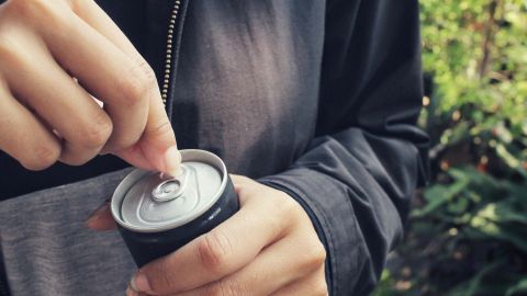 Eine Person öffnet eine Dose eines Energy-Drinks in einer grünen, naturnahen Umgebung.