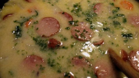 Eine cremige Suppe mit Stückchen von Cabanossi schwimmt in einer Schüssel, umgeben von frischen Kräutern und einer Holzlöffel.