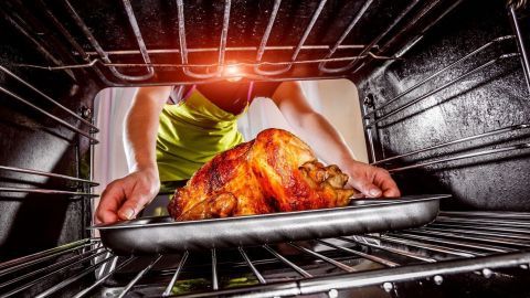 Eine Person zieht ein Blech mit einem goldbraun gebratenen Hähnchen aus dem Backofen, die Küche ist hell erleuchtet.