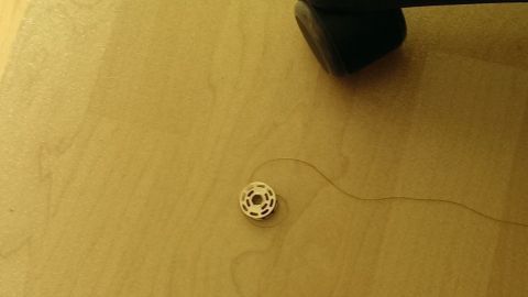 Ein Garnspulen-Objekt liegt auf dem Boden, während ein Stuhlbein in der Nähe steht, und ein Faden zieht sich davon weg.