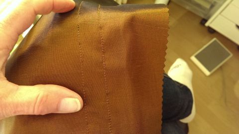 Eine Hand hält ein braunes Stück Stoff mit runder Naht über dem Knie, während die Person entspannt sitzt.