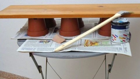 Eine Holzplatte ruht auf mehreren Plastiktöpfen und wird mit einer Farbrolle gestrichen, um einen Tisch zu schaffen.