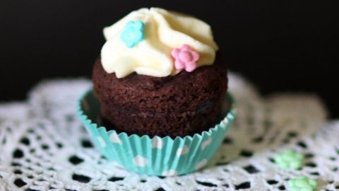 Ein schokoladiger Muffin mit Quarktopping steht auf einem weißen Spitzendeckchen und ist mit bunten Zuckerblümchen dekoriert.
