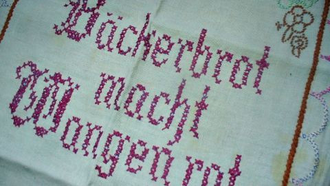 Ein handgesticktes, antikes Tischdeckchen zeigt den Spruch: „Bäckerbrot macht Wangen rot“ auf hellgrünem Stoff.