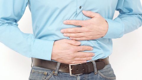 Ein Mann in einem blauen Hemd hält sich den Bauch, was auf Sodbrennen hindeutet, in einem neutralen Raum steht er da.