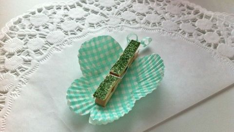 Eine grüne Muffinform ist dekorativ gefaltet und hält eine Holzklammer, die als Schmetterling dient, auf einem weißen Tisch.