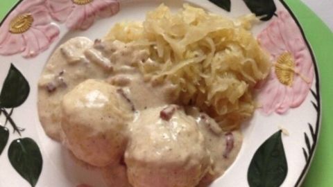 Eine garnierte Porzellanplatte zeigt Klöße in Speckrahmsoße neben Sauerkraut, angerichtet in einer gemütlichen Küche.