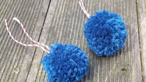 Zwei blaue PomPoms liegen auf einem Holzbrett, mit einer Schnur zum Aufhängen. Sie sind flauschig und handgemacht.