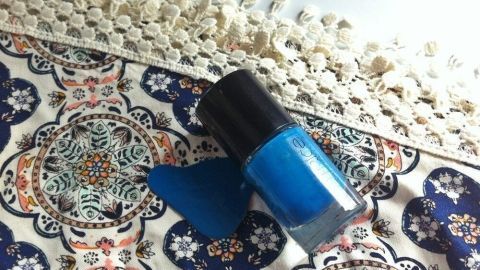 Ein blauer Nagellack steht auf einem bunten, gemusterten Stoff, daneben ein tropfenförmiger Nagellack-Fleck.