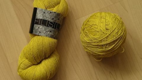 Ein gelber Wollstrang liegt neben einem gleichfarbigen Knäuel auf einem hellen Holztisch und wartet auf die nächste kreative Nutzung.
