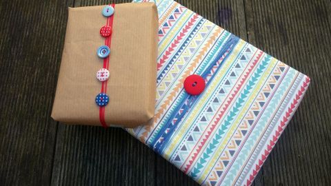 Zwei kreativ verpackte Geschenke mit bunten Knöpfen dekoriert, liegen auf einem rustikalen Holztisch.