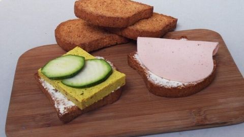 Zwieback liegt auf einem Holzbrett, zusammen mit belegtem Brot mit Gurken und Aufschnitt, vor neutralem Hintergrund.