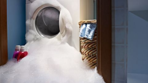 Eine Waschmaschine steht überflutet von Schaum, der den Boden des Waschraums bedeckt und einen Wäschekorb umgibt.