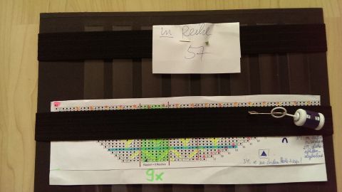 Ein Strickmusterblatt liegt auf einem Tisch, während eine Maschenprobe mit farbigen Markierungen darauf sichtbar ist.