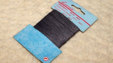 Stopfgarn auf einer blauen Verpackung zeigt die Möglichkeit, Knöpfe fest und elastisch anzunähen. Der Hintergrund ist beige.
