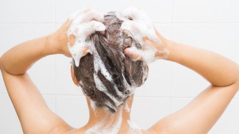 Eine Person mit schaumigem Shampoo in den Haaren massiert ihre Kopfhaut in einer hellen Badezimmerumgebung.