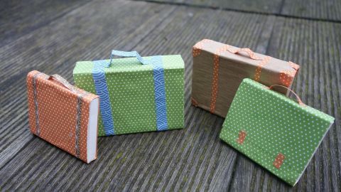 Bunte Minikoffer aus Papier stehen auf einem Holztisch, bereit für die Puppenstube und zum kreativen Spielen.