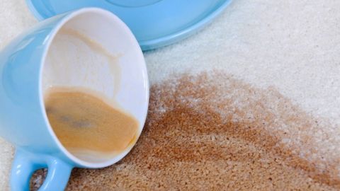 Eine umgefallene, hellblaue Tasse liegt auf einem hellen Teppich, während Kaffeeflecken sich ausbreiten.