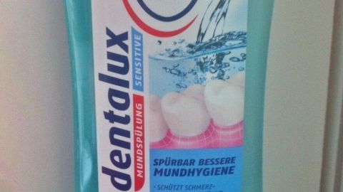Eine Flasche Zahnspülung mit der Aufschrift „dentalux Sensitiv“ steht auf einem hellen Hintergrund, umgeben von einer Wasserillustration.