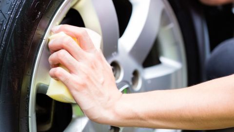 Eine Hand reinigt mit einem Schwamm aufmerksam eine Alufelge, während das Auto in einer hellen Umgebung steht.