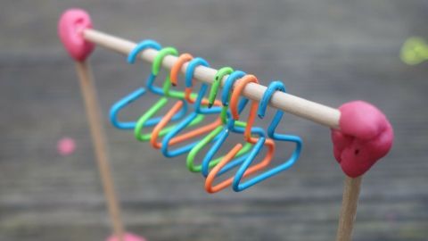Mini-Kleiderbügel hängen an einem stabilen Gestell aus Holzstäbchen, umgeben von buntem Knetmaterial auf einem Tisch.