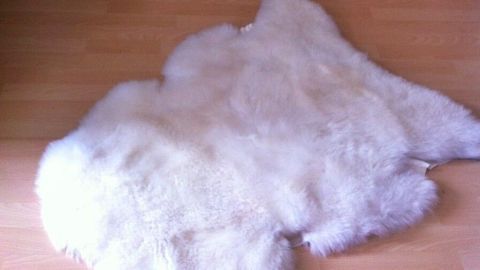 Ein weiches, helles Lammfell liegt auf einem Holzboden in einem gemütlichen Raum, umgeben von Kerzen und einer Laterne.