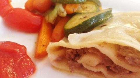 Eine Nudel-Hackfleischrolle liegt auf einem weißen Teller, begleitet von Ofengemüse und einer Tomatensauce.