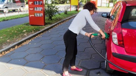 Eine Frau tankt an einer Zapfsäule, während im Hintergrund die Spritpreise angezeigt werden. Die Szene ist im Freien.