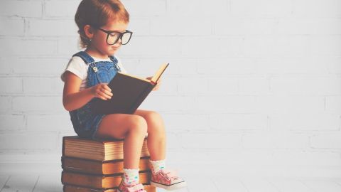 Ein Kind sitzt auf Büchern und liest ein Buch. Die Szene ist hell und schlicht mit einer weißen Wand im Hintergrund.