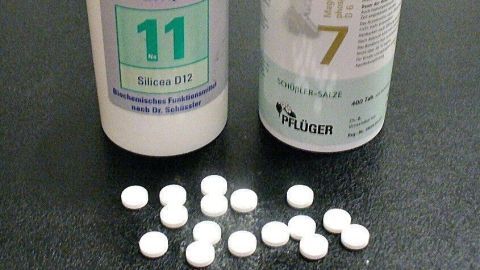 Zwei weißere Fläschchen mit Schüßler Salzen stehen nebeneinander, umgeben von kleinen weißen Tabletten auf einem dunklen Tisch.