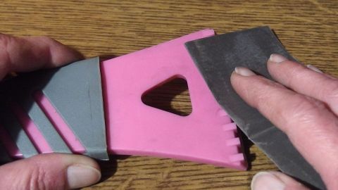 Alten Eiskratzer mit 600er Schleifpapier wieder schärfen. Ein Eiskratzer mit pinkem und grauem Design wird geschärft, während Hände den Schärfprozess auf einem Tisch durchführen.